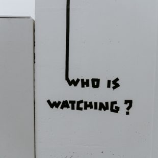 Tag mural who is watching ? en noir sur mur blanc.