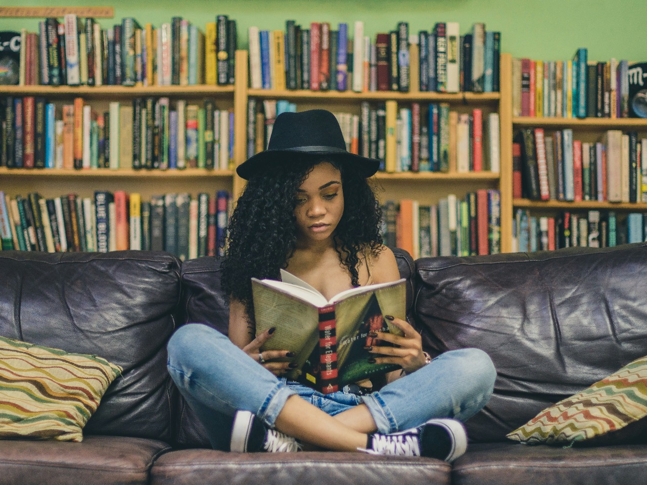 Femme assise sur un canapé en train de lire avec une bibliothèque en fond.