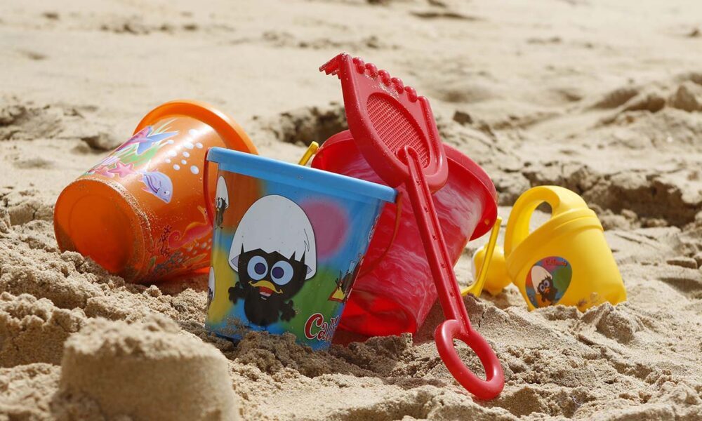 Photographie en paysage et en couleurs représentant en gros plan des jouets de plage en plastique sur du sable. On percoit les rayonnements du soleil.