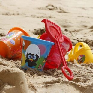 Photographie en paysage et en couleurs représentant en gros plan des jouets de plage en plastique sur du sable. On percoit les rayonnements du soleil.