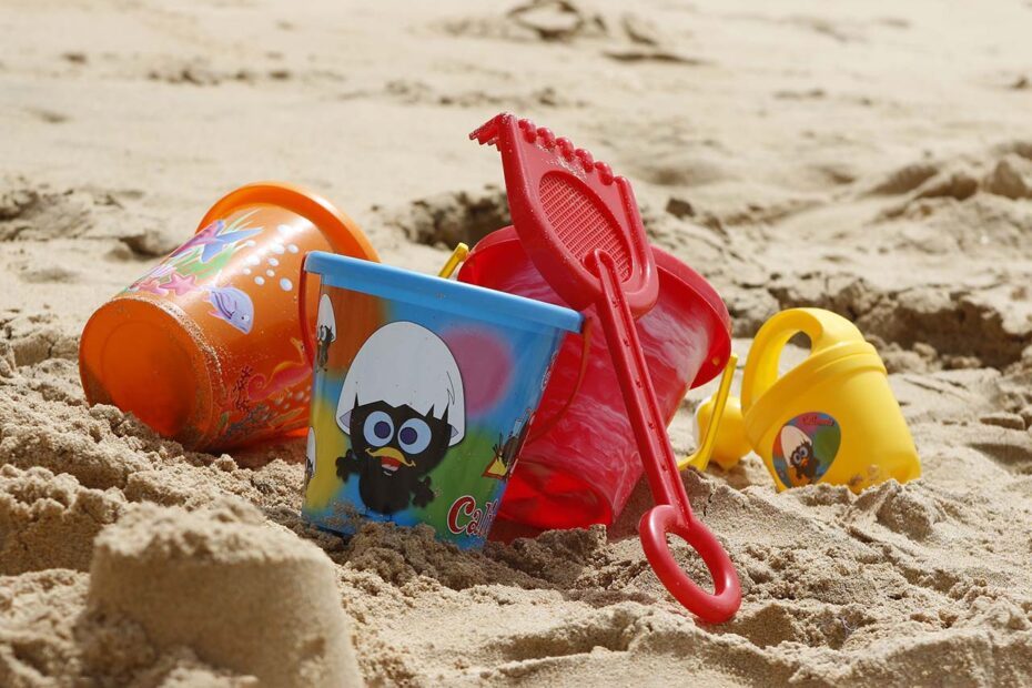 Photographie en paysage et en couleurs représentant en gros plan des jouets de plage en plastique sur du sable. On percoit les rayonnements du soleil.