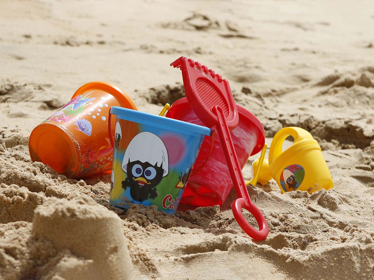 Photographie en paysage et en couleurs représentant en gros plan des jouets de plage en plastique sur du sable. On percoit les rayonnements du soleil.