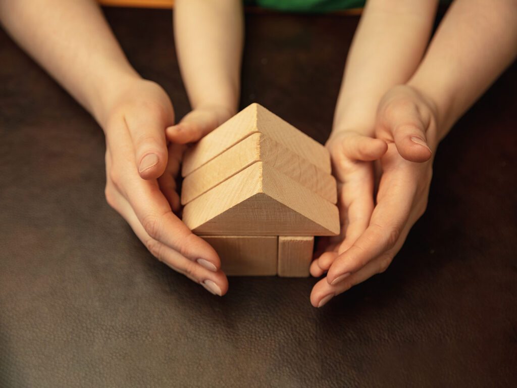 Mains d'adulte recouvrant des mains d'enfant et tenant ensemble une maison constituée de jeux en bois