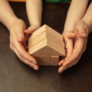 Mains d'adulte recouvrant des mains d'enfant et tenant ensemble une maison constituée de jeux en bois