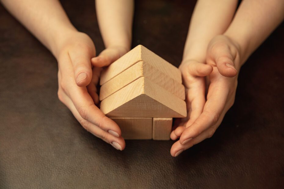 Mains d'adulte recouvrant des mains d'enfant et tenant ensemble une maison constituée de jeux en bois