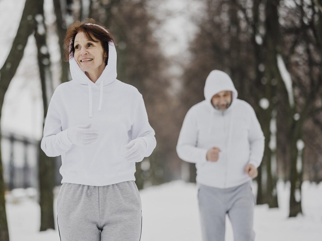 Photographie en couleurs de format paysage montrant une femme au premier plan et un homme au second plan dehors dans un paysage de neige et d'arbres. Les 2 personnes sont en train de faire leur footing. L'ambiance est hivernale avec une lumière très blanche.