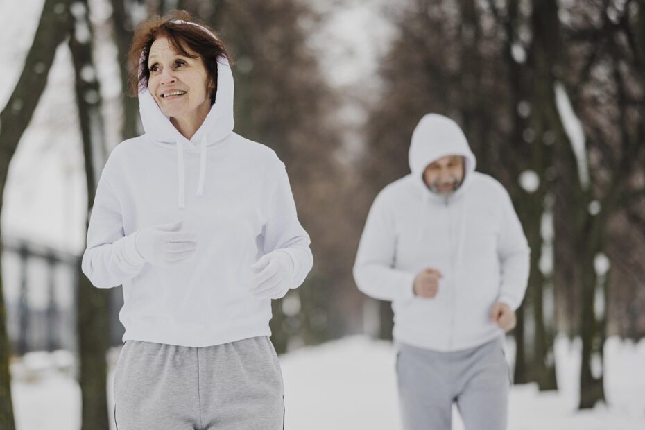 Photographie en couleurs de format paysage montrant une femme au premier plan et un homme au second plan dehors dans un paysage de neige et d'arbres. Les 2 personnes sont en train de faire leur footing. L'ambiance est hivernale avec une lumière très blanche.