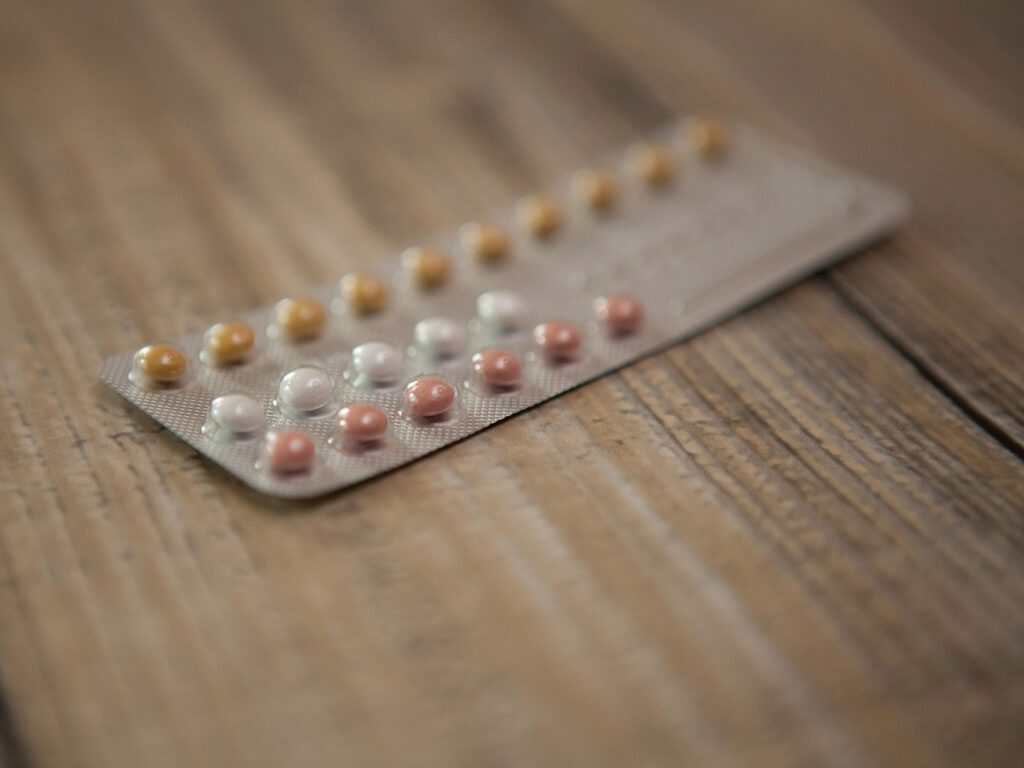 Photographie en gros plan d'une plaquette de pilules contraceptive posée sur un meuble en bois. Le point est fait sur quelques pilules colorées en jaune, blanc et rose, et le reste de la photo est flou. L'image est en couleurs.