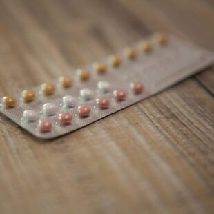 Photographie en gros plan d'une plaquette de pilules contraceptive posée sur un meuble en bois. Le point est fait sur quelques pilules colorées en jaune, blanc et rose, et le reste de la photo est flou. L'image est en couleurs.