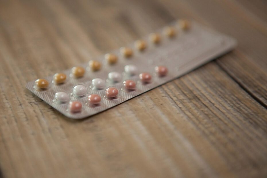 Photographie en gros plan d'une plaquette de pilules contraceptive posée sur un meuble en bois. Le point est fait sur quelques pilules colorées en jaune, blanc et rose, et le reste de la photo est flou. L'image est en couleurs.