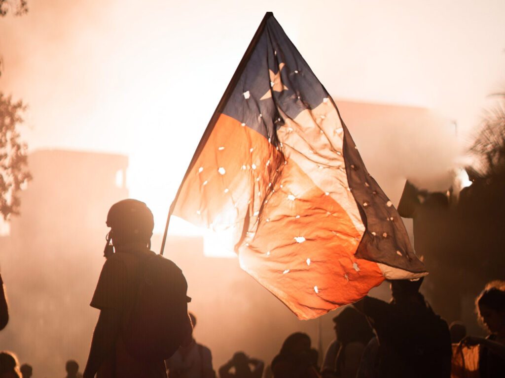 Photographie en paysage et en couleurs d'une scène de manifestation au Chili. Le premier plan montre un manifestant portant le drapeau chilien cribblé d'impacts de balles. La prise de vue est en lègère contre-plongée, laissant apparaître le ciel dans lequel se détache le drapeau au centre de l'image, et une lumière laissant supposer la présence de fumigènes. La foule apparaît au second plan.